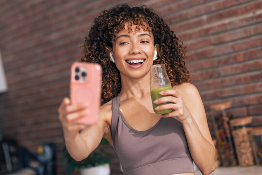 Smiling sporty woman taking selfie with smartphone while drinking detox juice at home