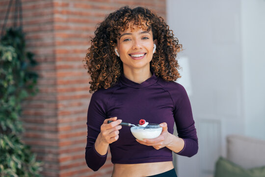 Smiling pretty woman eating healthy yogurt bowl while looking at camera at home