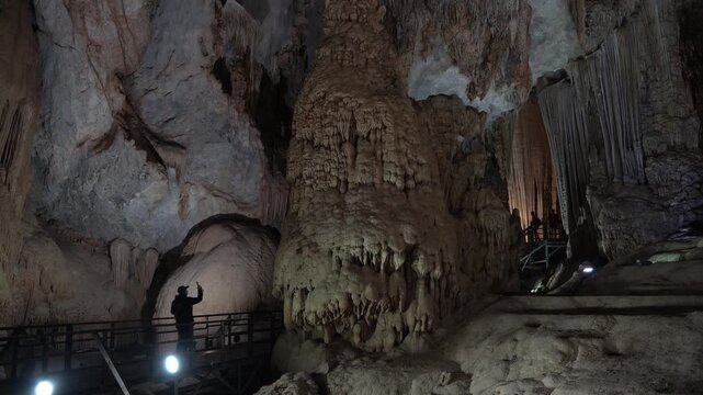 Tourist taking a selfie on a wooden pathway inside a majestic, illuminated cavern with stalactites