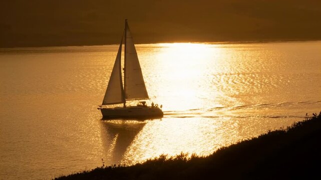 Sailing sloop with backlit crew moving right across sunset bay on outing, breeze filling mainsail