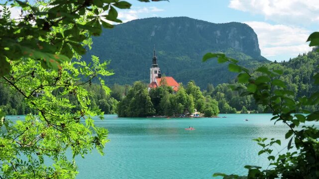 View of the iconic Church of the Assumption on Bled Island in Lake Bled, Slovenia, framed by green tree leaves on a sunny summer day with crystal-clear alpine scenery.