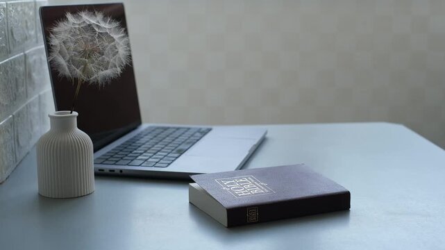 Dandelion in vase and Holy Bible beside laptop on desk
