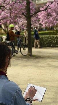 An artist sits outdoors and draws in a spiral notebook. The background shows a public park with blooming trees, a bicycle, and people walking by.