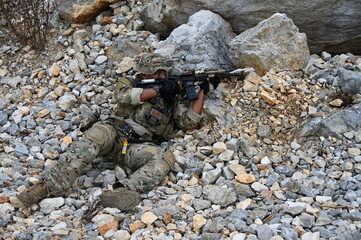 Obraz premium Focused military infantry soldier lying prone on rugged rocky terrain aiming a tactical assault rifle during a dangerous combat training mission for national defense and special security.