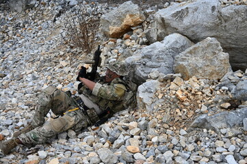 Obraz premium Focused military infantry soldier lying prone on rugged rocky terrain aiming a tactical assault rifle during a dangerous combat training mission for national defense and special security.