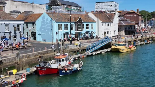 Weymouth Dorset England UK. 08.07.2026. Video, Boats bob gently in the water next to a waterfront street lined with colourful buildings and people enjoying outdoor seating