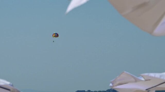 A brightly colored tandem parasail flies high against a clear blue sky over a distant coastline, framed by out-of-focus white beach umbrellas in the foreground.