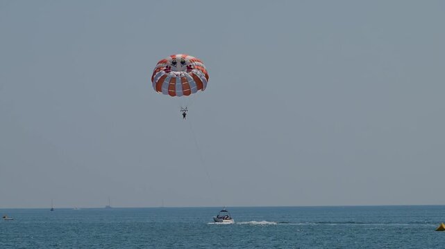A red and white parasail carrying two people is towed by a motorboat across a calm, clear blue sea under a cloudless sky.
