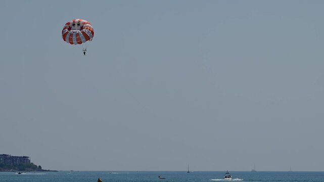 Two people enjoy a thrilling tandem parasailing ride under a red and white striped parachute, towed by a speedboat across a calm blue sea on a clear summer day.