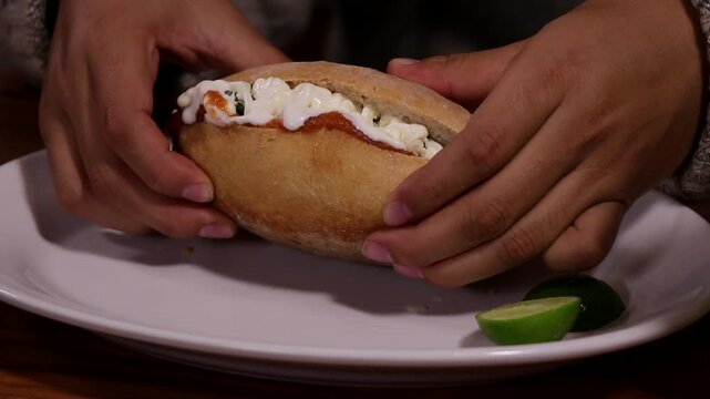 Mujer tomando una torta de chilaquiles lista para comer. Torta de chilaquiles servida en plato blanco. Torta de chilaquiles con crema y queso fresco. Comida callejera mexicana capturada en detalle