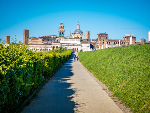 People walking on a clear path lined by green hedges and grass along a park, approaching the historic skyline of Mantova, Italy, with its distinctive bell towers and domes