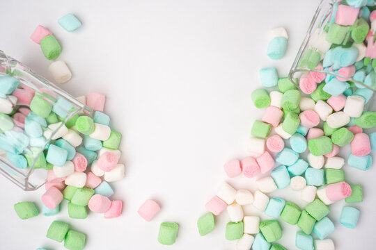 Pastel pink, green, blue, and white mini marshmallows spill from two clear glass jars onto a clean white background. They scatter across the surface, forming a sweet and colorful treat scene