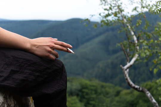 Close up view of a person holding a burning cigarette in their hand while sitting on a viewpoint with lush green hills and forest background.
