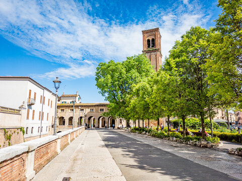 Mantova. Italy. Displaying an urban square with an ancient bell tower. Historical architecture featuring arched porticos. And vibrant green trees under a clear sky