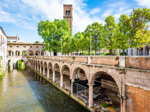 Mantova, Italy, featuring the historic Pescherie di Giulio Romano with its arcades alongside the Rio Canal, showing classic Italian architecture and a distant bell tower under a bright sky