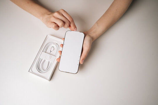Top view of woman unpacking new smartphone and removing screen protector sitting on white table, with charging cable in box visible in background. Concept of excitement of unboxing new device.