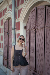 Obraz premium Woman smiles near antique red wooden gates on sunny Moscow street. Urban portrait, travel, heritage architecture, fashion, lifestyle, confidence, summer
