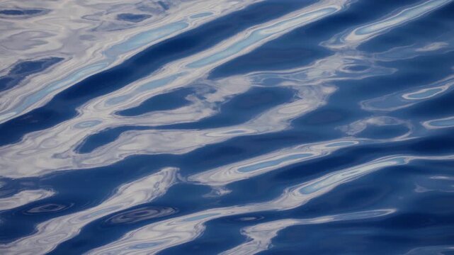 Serene blue ocean water surface with gentle rolling ripples creating abstract patterns. Soft white and turquoise sky reflections dance across the calm sea in a mesmerizing display.