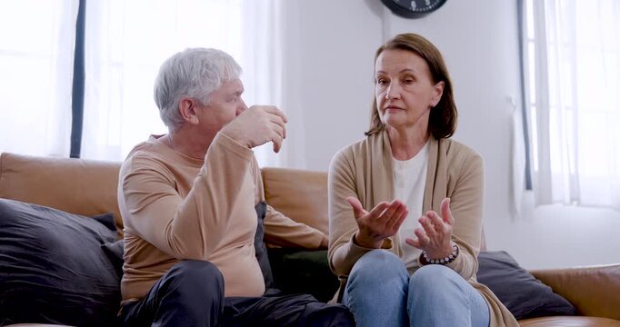 Mature Caucasian husband and wife sit face to face on sofa, gesture with hands, listen, and respond during thoughtful conversation built on trust, communication, emotional support, and respect.