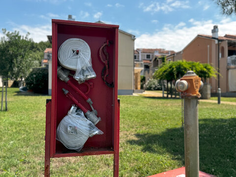 Fire safety equipment set with rolled hoses and nozzles in a red metal box outdoors. Residential area fire fighting station, street protection hydrant and tools cabinet near residential buildings