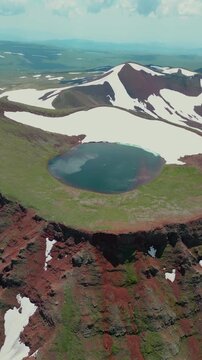 Vertical Drone Shot of Volcanic Crater Lake surrounded by Red Soil, Green Slopes, and Melting Snow
