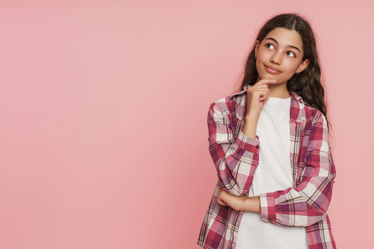Thoughtful mixed race schoolgirl smiling and looking at copy space with hand on chin on pink studio background, human emotions, imagination and childhood expression concept