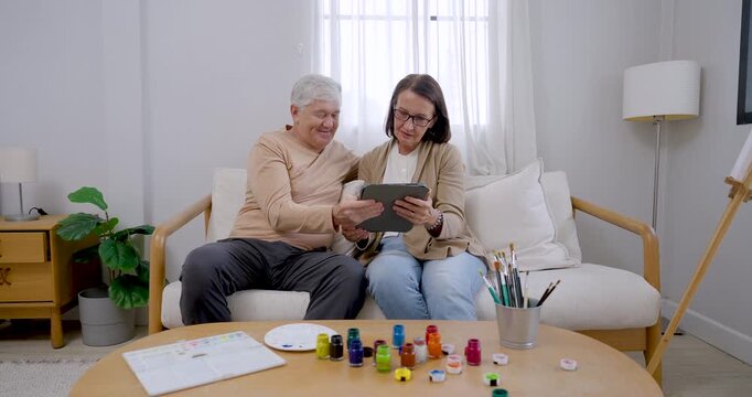 Senior husband and wife sit near paint supplies, pointing at tablet and selecting reference before painting, sharing cheerful conversation, creative inspiration, bonding, and retirement wellness.