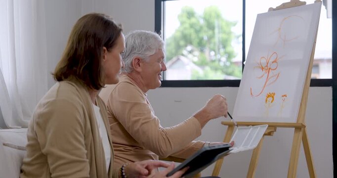 Senior man adds flower details to canvas, then checks tablet with senior woman beside him, creating home art therapy moment supporting emotional wellness, mindful relaxation, active retirement.