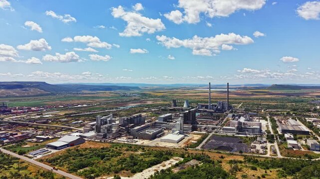 A wide view of an industrial area showing factories and power plants surrounded by fields and hills. The sky is bright with a few clouds, creating a sunny atmosphere.