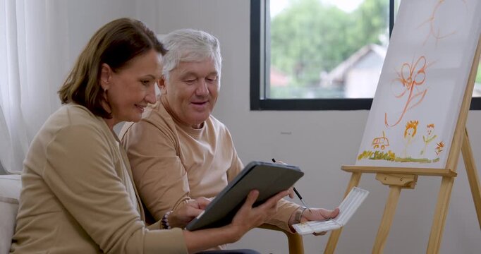 Senior man paints flower artwork, pauses to view tablet with senior woman, then returns brush to canvas, sharing home art therapy activity supporting calm emotion, companionship, healthy aging.