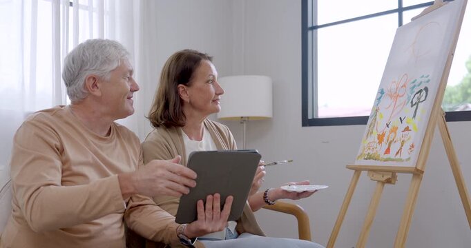 Senior couple compare tablet reference with canvas while woman holds brush and palette, pausing to discuss ideas and support cognitive stimulation, companionship, and creative wellness at home.