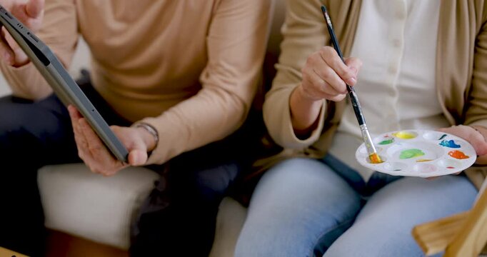 Senior woman mixes paint on palette and lifts brush while senior husband checks tablet nearby, sharing home art therapy for creativity, cognitive stimulation, companionship, active retirement.