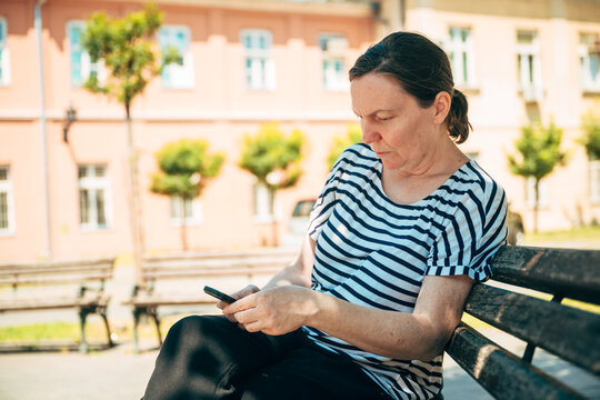 Middle-aged woman using a smartphone while sitting on a shaded park bench in summer. Mobile technology, outdoor lifestyle and digital communication concept.