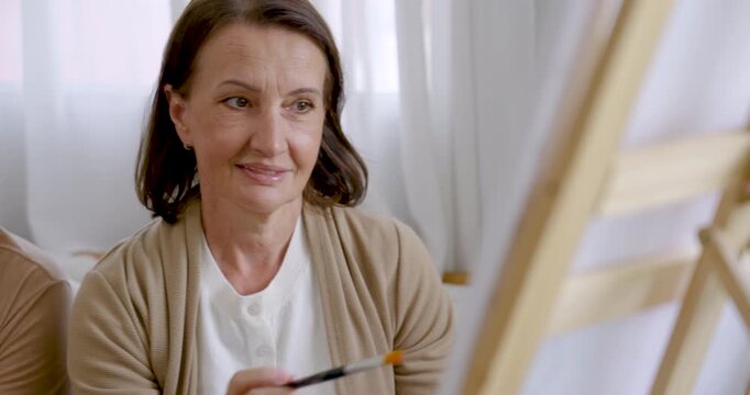 Senior woman holds paintbrush, talks, and smiles while looking toward canvas during art therapy at home, expressing creative confidence, mindful relaxation, emotional wellness, and active retirement.