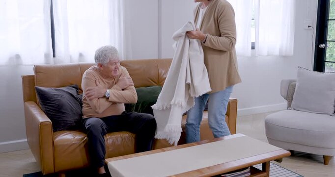 Senior Caucasian woman approaches husband resting on sofa, wraps blanket across shoulders, sits beside him, and checks comfort with caring touch during supportive retirement life at home together.