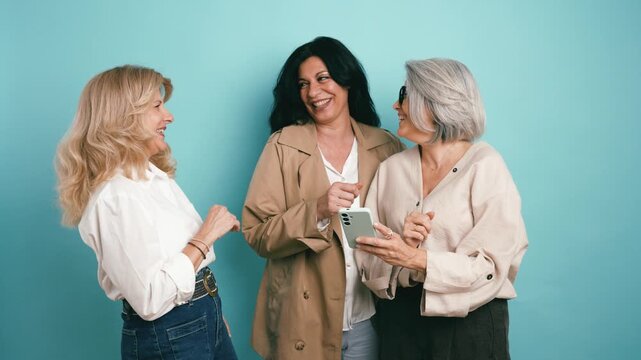 Group of happy mature women friends looking at a mobile phone screen, laughing and chatting together against a solid blue background