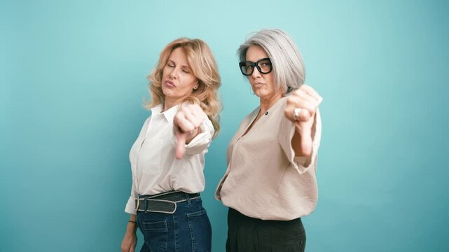 Two angry and disappointed businesswomen expressing negativity. Unhappy senior women making a thumbs down gesture on a turquoise background