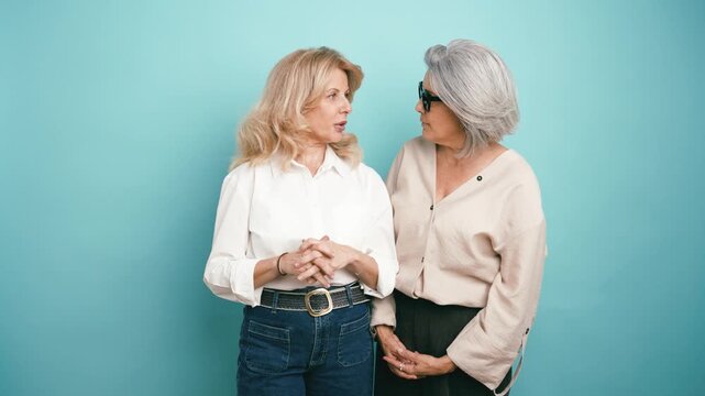 Two stylish mature women having an engaging conversation, gesturing and expressing emotions while standing against a solid blue background