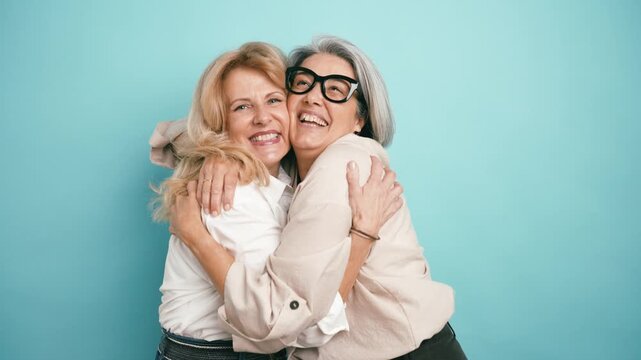 Two smiling elderly women friends hugging each other with genuine affection. Happy mature female friendship embracing on a blue background