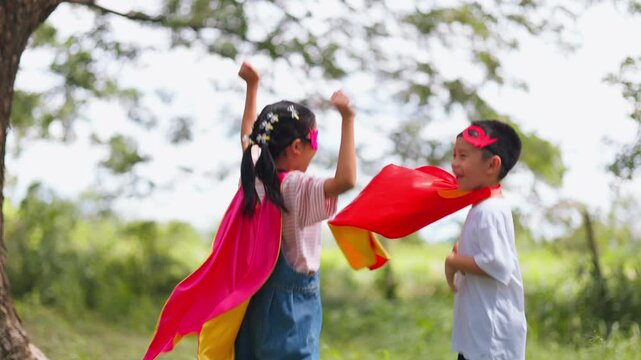 Active Asian children wearing superhero cape and mask having fun running in tree tunnel park. Kids imagination, creative future dream, and brave hero inspiration activity.