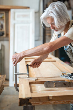 Talented man restorer in woodworking shop fixating antique wooden door at workbench using clamps, professional craftsman preparing object for reconstruction process at handmade carpentry workshop.
