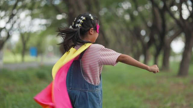 Active Asian children wearing superhero cape and mask having fun running in tree tunnel park. Kids imagination, creative future dream, and brave hero inspiration activity.
