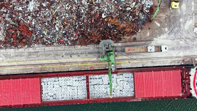 A cargo ship is being loaded with metal scraps at a busy port. The ship is parked near a large pile of metal debris. Cranes are operating to move materials efficiently.