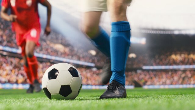 A blue-team striker prepares a free kick and focuses on technique before driving the ball toward the net, captured from a low angle during an attacking set piece in live action on the soccer pitch.