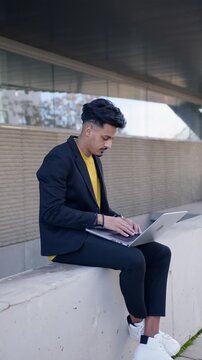 Young moroccan businessman working remotely on his laptop. Digital nomad typing on the computer keyboard while sitting outdoors in the city. Vertical