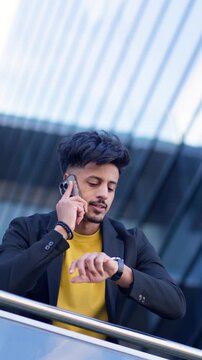 Young moroccan businessman talking on a smartphone and checking his watch. An arab professional having a business conversation outdoors. Vertical