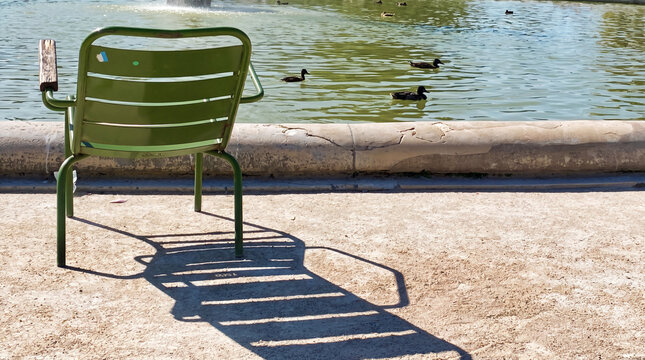 A solitary green metal chair facing a tranquil lake with ducks swimming in the background. The shadow and calm waterfront create a peaceful atmosphere, symbolizing relaxation, solitude