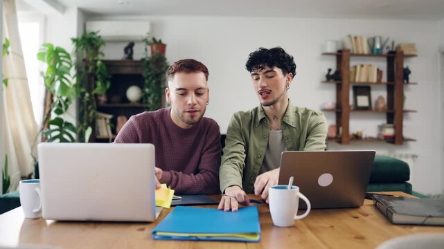 Two young gay men collaborating on a business project at home. They are analyzing documents and discussing ideas while working on their laptops