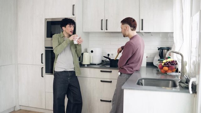 Happy male couple spending quality time together in the morning. One man is cooking on the stove while his partner drinks a hot beverage and talks