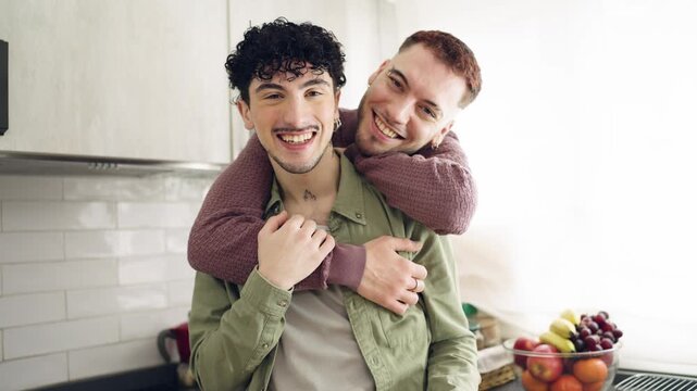 Happy young gay couple sharing a loving moment at home. One man tenderly hugs his boyfriend from behind while both smile at the camera